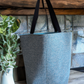 Textured blue tote bag with black straps on a wooden surface next to a plant.