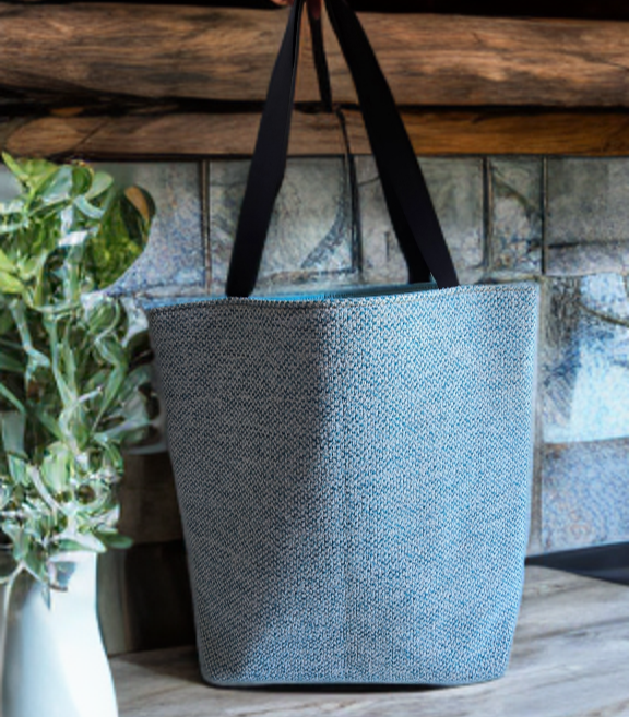 Textured blue tote bag with black straps on a wooden surface next to a plant.